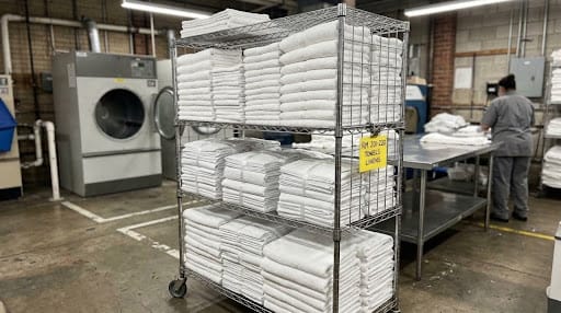 Stacks of professionally laundered white hotel linens and towels organized on a rolling wire rack inside a commercial laundry facility.